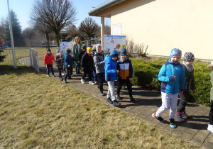 Dzieci wraz z panią maszerują z wiosennymi plakatami pod budynkiem przedszkola.